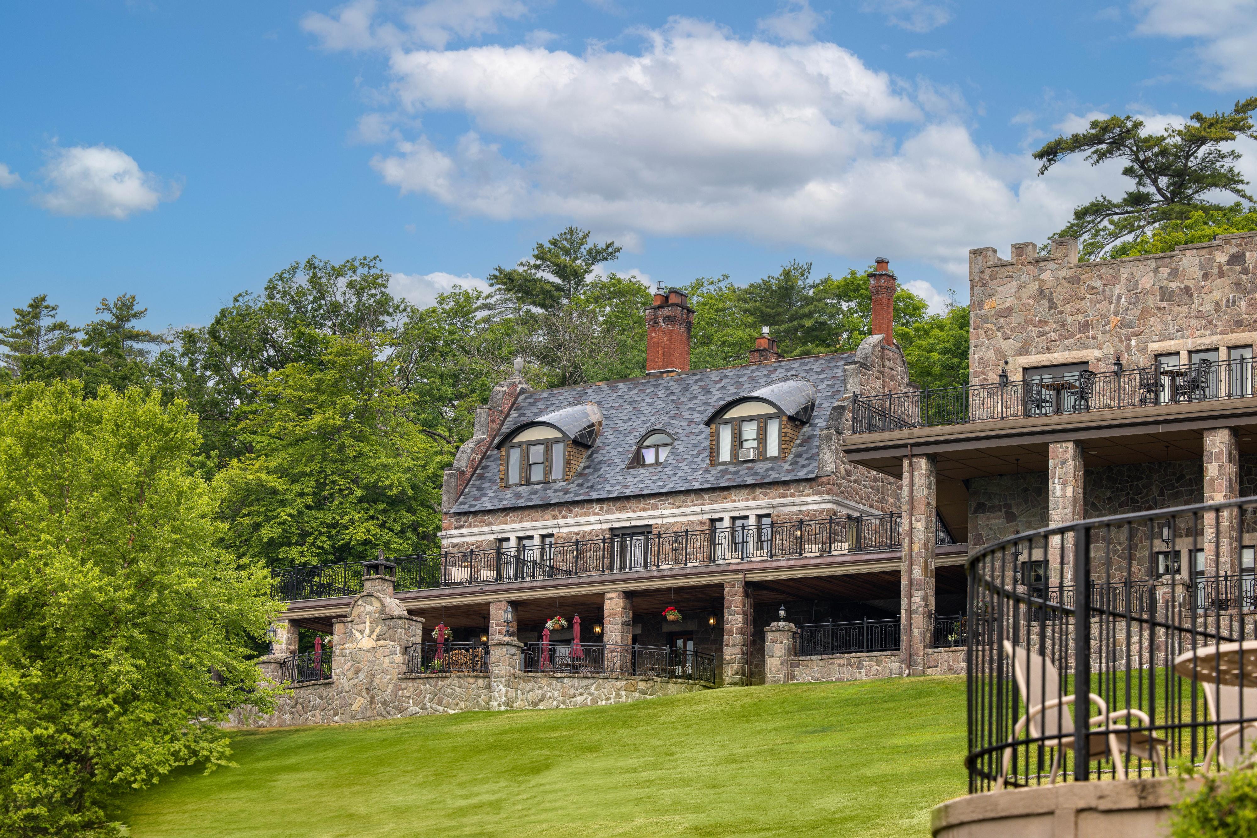 A large stone building with a porch and balcony, surrounded by trees and a green lawn.