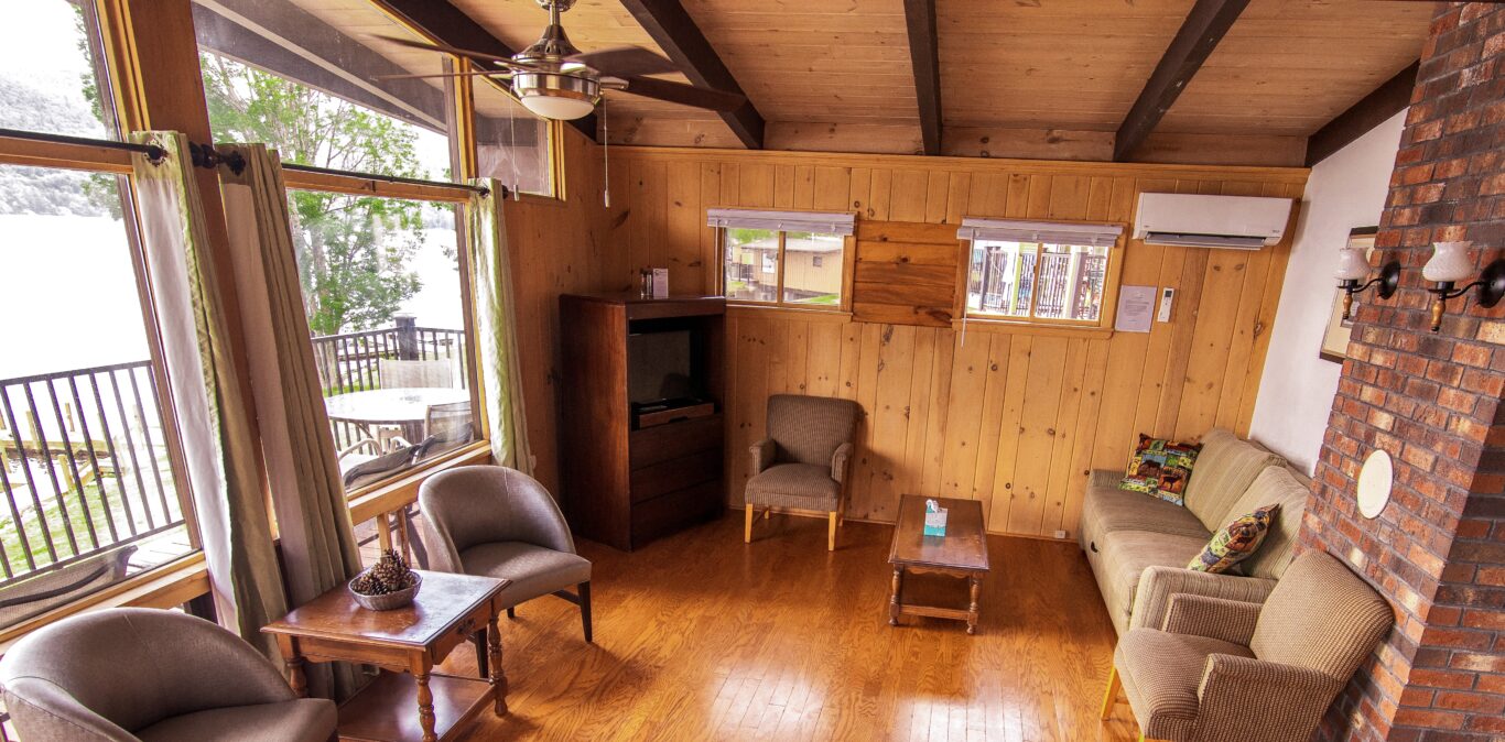 A cozy living room with wood paneling, a brick fireplace, and large windows overlooking a deck.