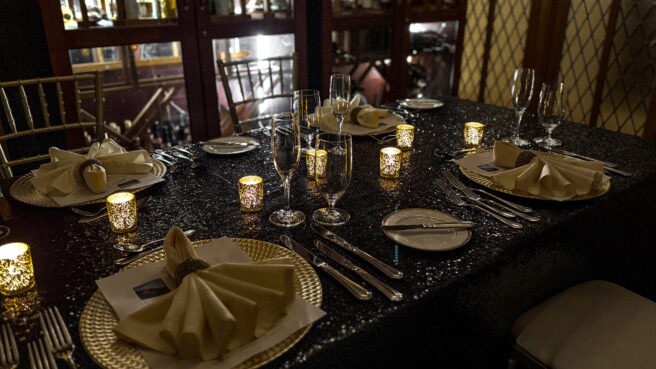 A luxurious and intimate formal dinner setting featuring a long table draped in a glittering black sequin cloth. The table is illuminated by a row of glowing gold mercury glass votives, creating a warm, amber ambiance.