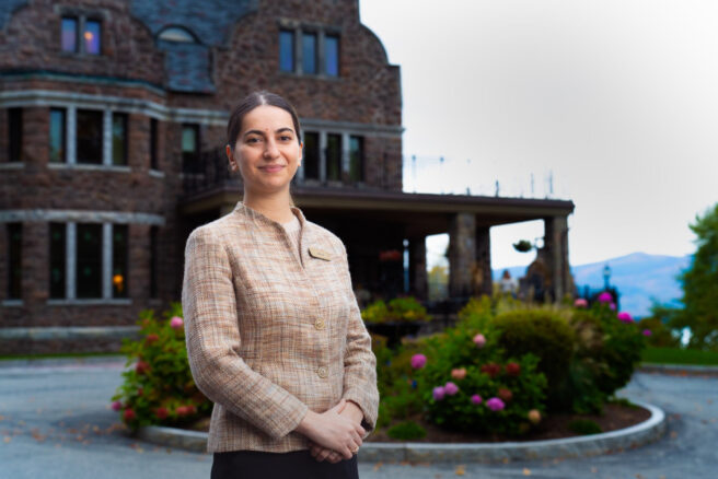 A woman stands outside the grand stone building of Erlowest, with lush bushes in the foreground and majestic mountains in the background, ready to meet with the friendly staff inside.