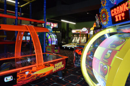 An arcade with vibrant games and machines lining the lanes, casting colorful glows in a dimly lit room.