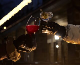 Two people toast with drinks in gloved hands by a fire pit at the Winterbar, one holding red wine and the other a cocktail garnished with an olive, all set against a backdrop of festive lights near Erlowest.