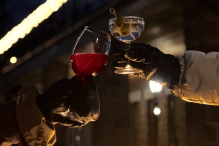 Two people toast with drinks in gloved hands by a fire pit at the Winterbar, one holding red wine and the other a cocktail garnished with an olive, all set against a backdrop of festive lights near Erlowest.