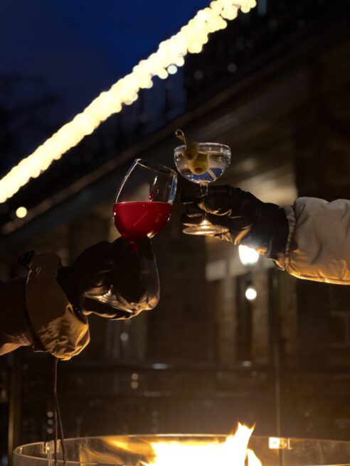 Two people toast with drinks in gloved hands by a fire pit at the Winterbar, one holding red wine and the other a cocktail garnished with an olive, all set against a backdrop of festive lights near Erlowest.