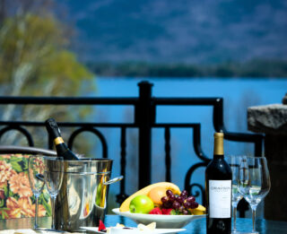 A patio table set for a Mother's Day brunch buffet, with wine, fruit, and cheese, overlooks a lake and mountains under a clear blue sky. Two chairs and two wine glasses invite you to celebrate in style.