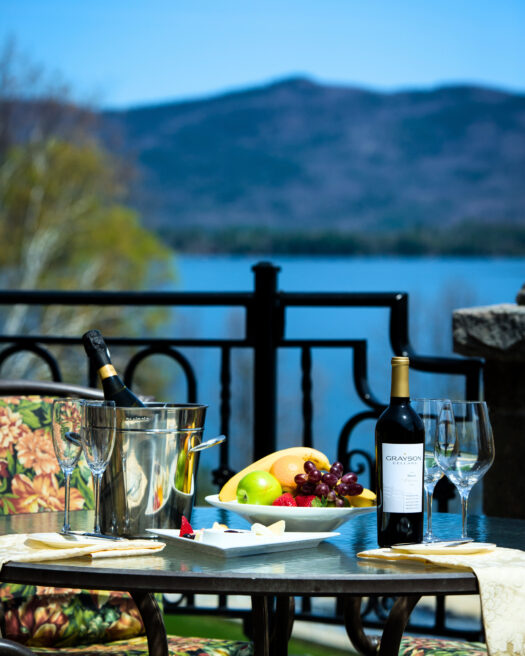 A patio table set for a Mother's Day brunch buffet, with wine, fruit, and cheese, overlooks a lake and mountains under a clear blue sky. Two chairs and two wine glasses invite you to celebrate in style.