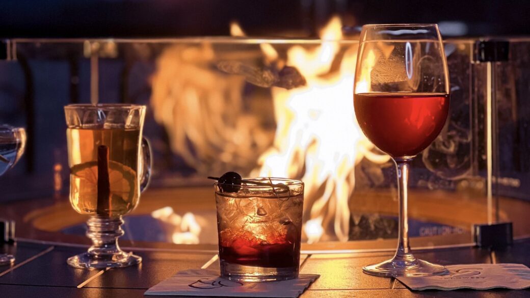 Three cocktails on a fire table with flames behind glass, overlooking Lake George