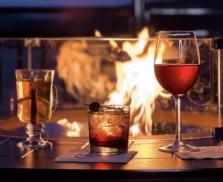 Three cocktails on a fire table with flames behind glass, overlooking Lake George