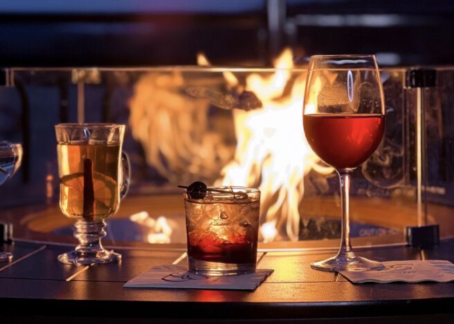Three cocktails on a fire table with flames behind glass, overlooking Lake George