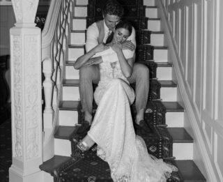 A bride and groom sit on a staircase in wedding attire, smiling and embracing each other in a black-and-white photo at one of the stunning Lake George luxury hotels.