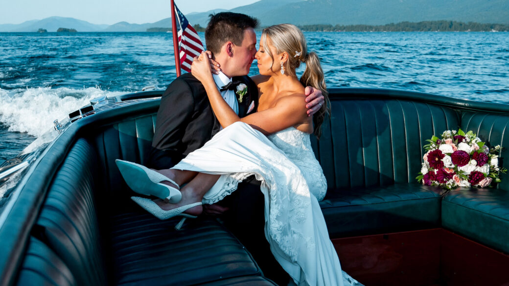 A bride and groom sit closely together on a boat near lake george luxury hotels, with the bride's bouquet beside them and an American flag at the back, surrounded by water and distant mountains.