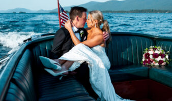 A bride and groom sit closely together on a boat near lake george luxury hotels, with the bride's bouquet beside them and an American flag at the back, surrounded by water and distant mountains.