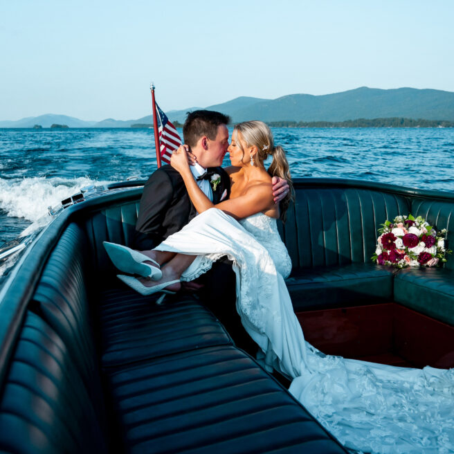 A bride and groom sit closely together on a boat near lake george luxury hotels, with the bride's bouquet beside them and an American flag at the back, surrounded by water and distant mountains.
