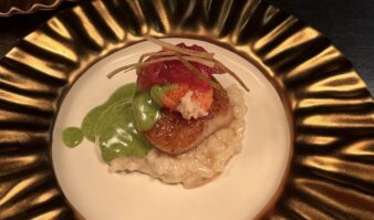 Seared scallop served over creamy risotto, topped with lobster and pickled garnish, with green herb sauce on a gold-rimmed plate
