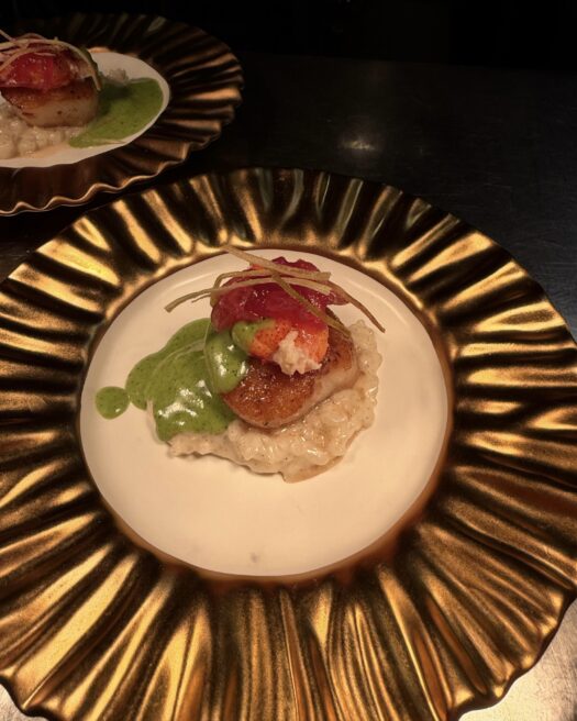 Seared scallop served over creamy risotto, topped with lobster and pickled garnish, with green herb sauce on a gold-rimmed plate