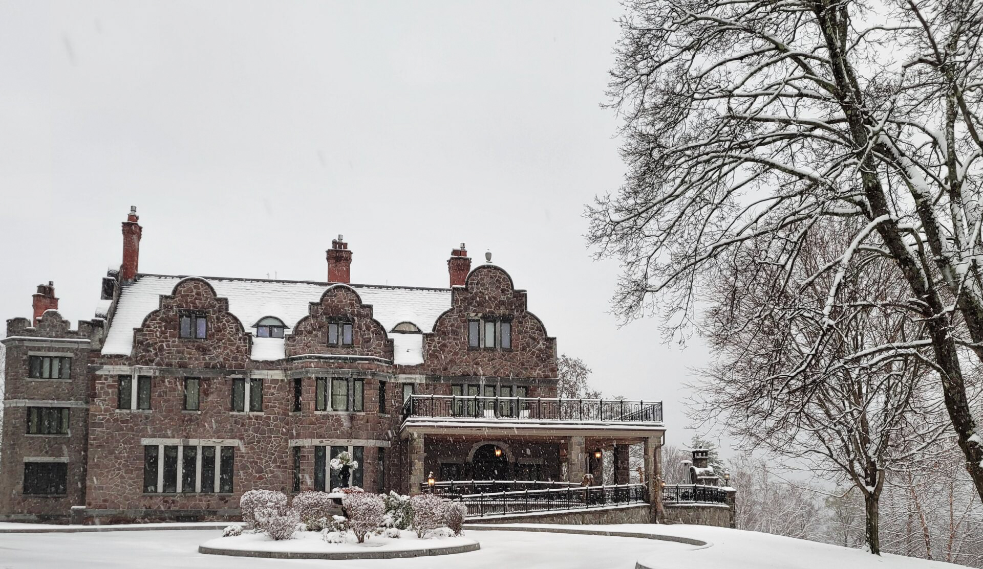 A large stone building with multiple chimneys and gabled roofs is surrounded by snow-covered trees and shrubs on a winter day.