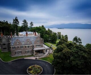 The Erlowest is a large stone mansion with multiple chimneys near a lake, surrounded by trees and mountains in the background under a cloudy sky. Terms and Conditions may apply for tours or stays at this historic site.