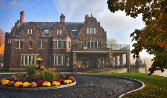 Large stone mansion with multiple chimneys, arched windows, and a circular driveway featuring a flowerbed, this impressive lodging is surrounded by autumn trees—offering a stunning stay near Lake George.
